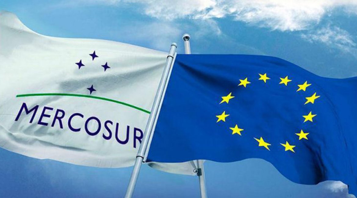 #pracegover duas bandeiras balançam ao vento altas em um céu azul, uma branca com a imagem de quatro estrelas representando o cruzeiro do sul, o símbolo do mercosul com a própria escrita abaixo, a outra azul com as estrelas amarelas em círculo, símbolo da união europeia.